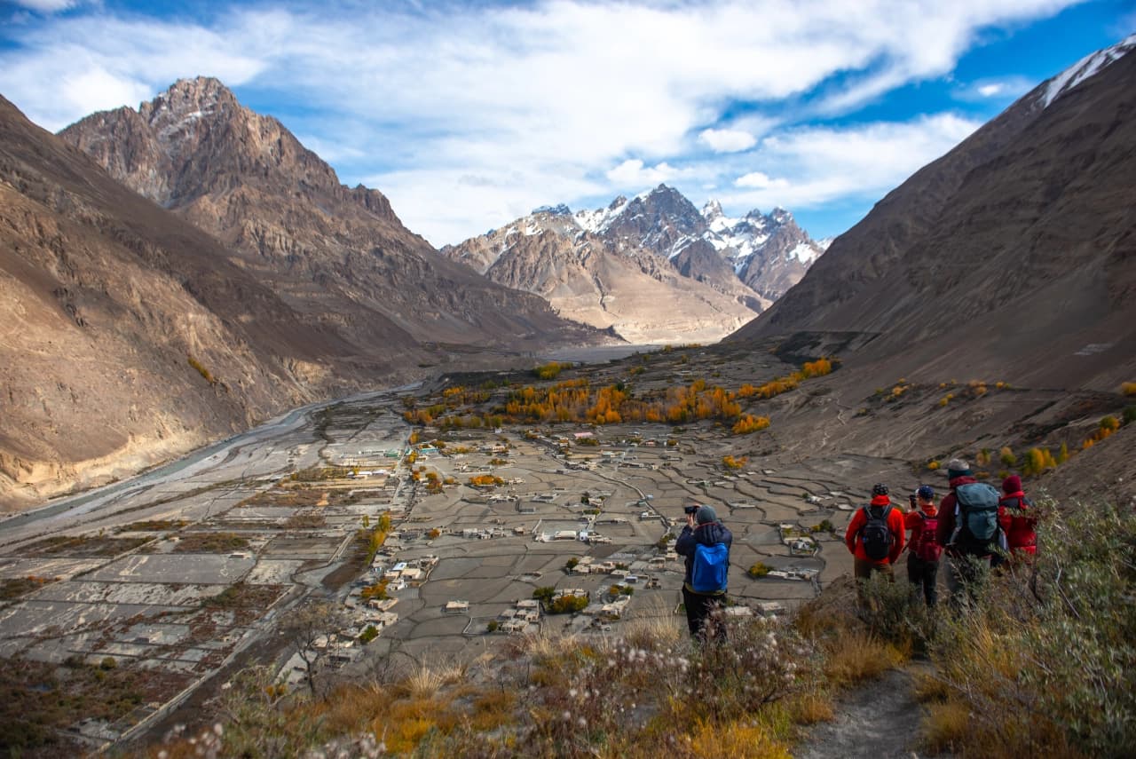 Hikers on a ridge above a mountain village, snow peaks, and autumn trees in northern Pakistan